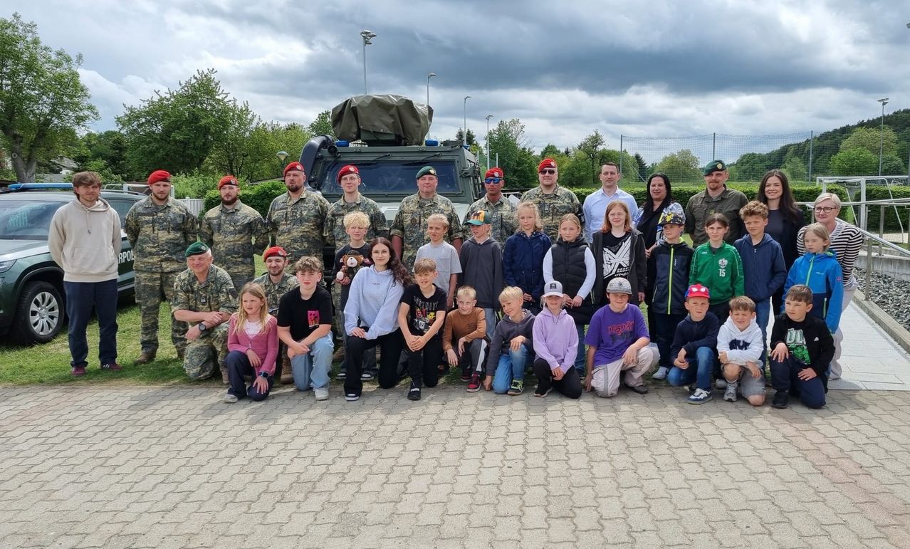 A group of adults and children pose for a photo in front of an armored vehicle. They wear military uniforms and hats, with some holding toys. Trees and streetlights are in the background.