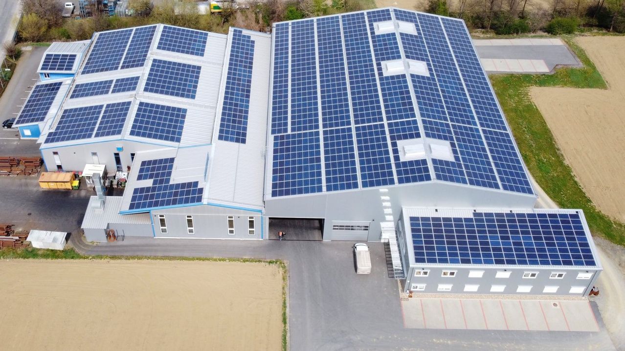 Luftaufnahme einer Fabrik mit zahlreichen Solarpanelen auf dem Dach und einem weißen LKW vor dem Gebäude.