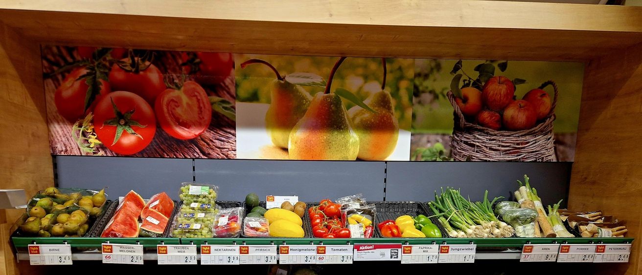 Ein Lebensmittelgeschäft mit verschiedenen Obst- und Gemüsesorten. Grüne Regale halten Tomaten, Orangen, Äpfel, Bananen, Birnen und mehr. An der Wand befindet sich ein großes Wandbild von Obst und Gemüse.