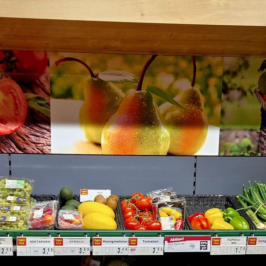 Ein Lebensmittelgeschäft mit verschiedenen Obst- und Gemüsesorten. Grüne Regale halten Tomaten, Orangen, Äpfel, Bananen, Birnen und mehr. An der Wand befindet sich ein großes Wandbild von Obst und Gemüse.