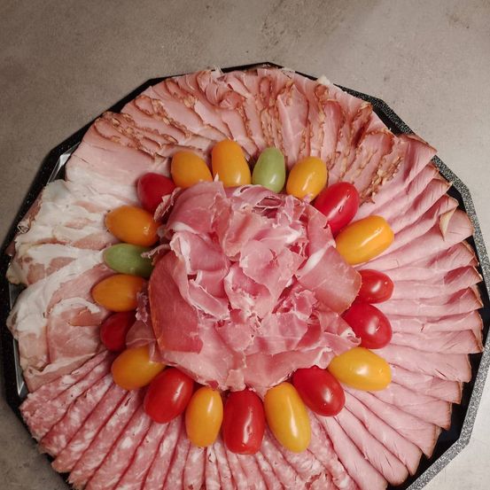 Eine runde Charcuterie-Platte mit verschiedenen rosa Fleischsorten, darunter geschnittener Schinken, umgeben von bunten Kirschtomaten. Die Platte steht auf einer grauen Oberfläche.