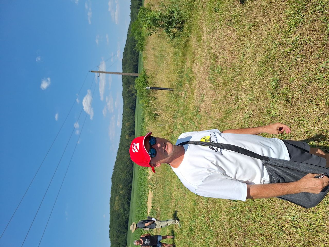 Ein Mann mit roter Kappe posiert für ein Foto in einem Grasfeld mit zwei anderen dahinter. Der Himmel ist klar mit einigen Wolken. Bäume und ein Mast mit Drähten sind im Hintergrund sichtbar.