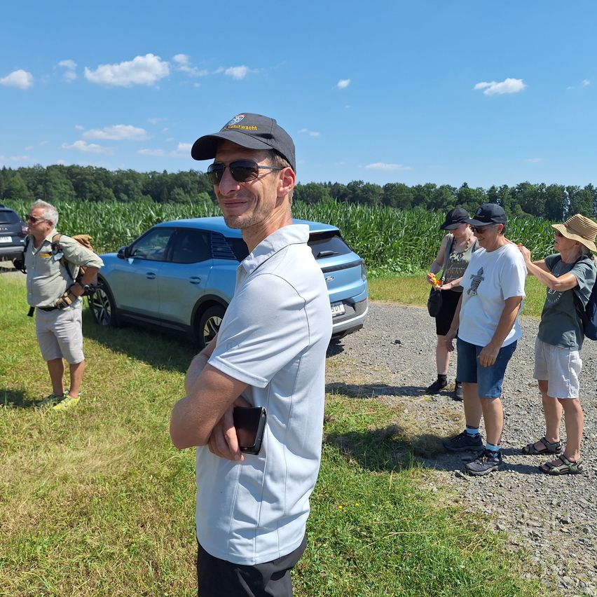Eine Gruppe von Menschen steht in einem Grasbereich in der Nähe eines Autos. Eine Person hält ein Telefon.