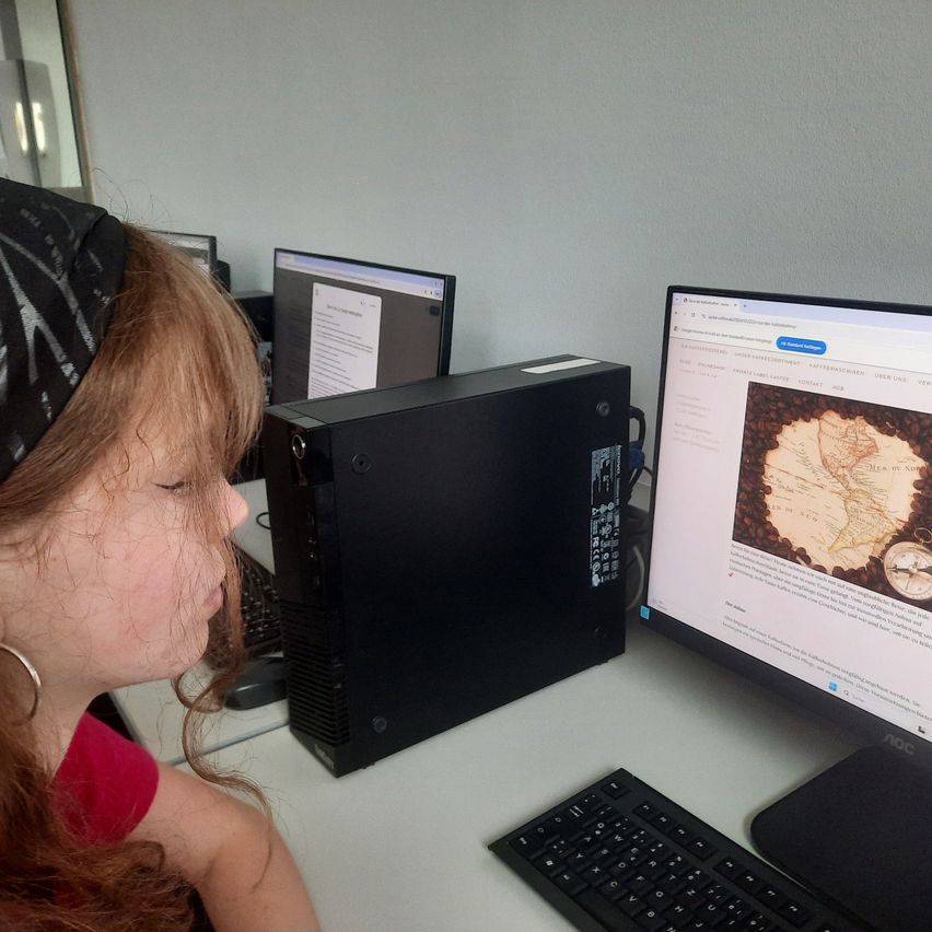 Eine junge Frau mit langen roten Haaren und einem schwarzen Kopftuch schaut auf einen Computerbildschirm, der eine Karte anzeigt. Der Computer steht auf einem Schreibtisch mit einer Tastatur und einem schwarzen Computerturm.