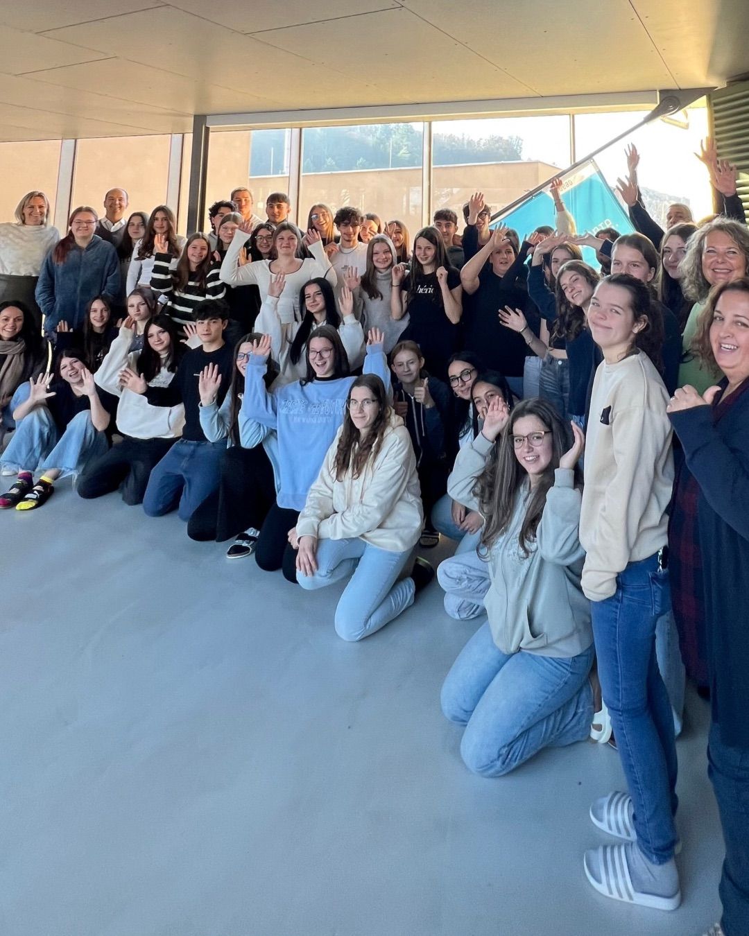 Eine Gruppe von Menschen posiert für ein Foto in einem Raum mit Glasfenstern. Sie tragen Freizeitkleidung. Einige sitzen, andere stehen. Alle lächeln und heben ihre Hände. Der Raum hat einen weißen Boden und wird von Sonnenlicht beleuchtet. Außerhalb des Raumes gibt es Bäume und Gebäude.