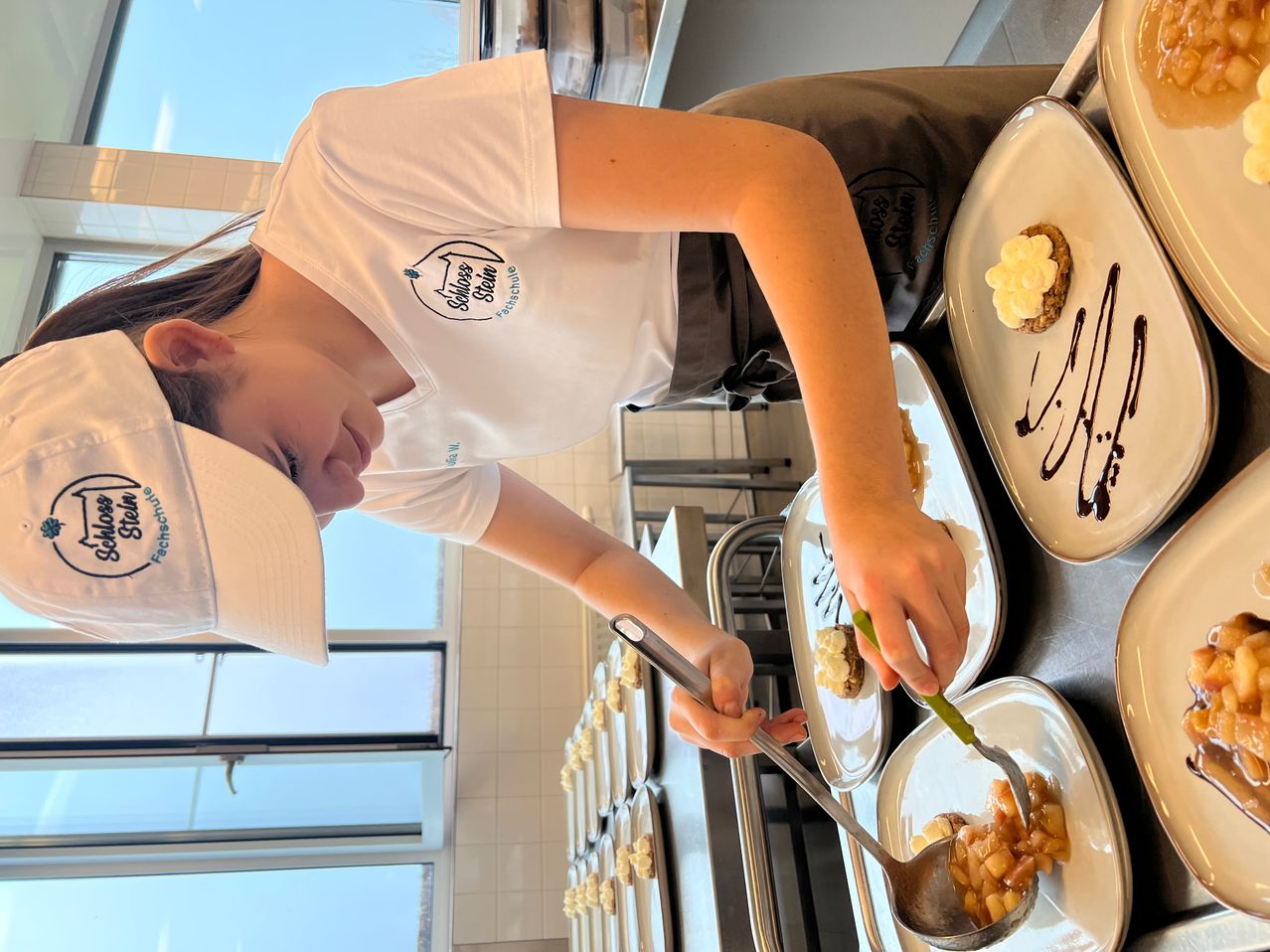 Eine Frau in einem weißen Shirt und einer Mütze ist in einer Küche und serviert Essen mit einem Löffel. Der Tisch hat mehrere Teller mit Desserts. Hinter ihr ist ein Fenster.