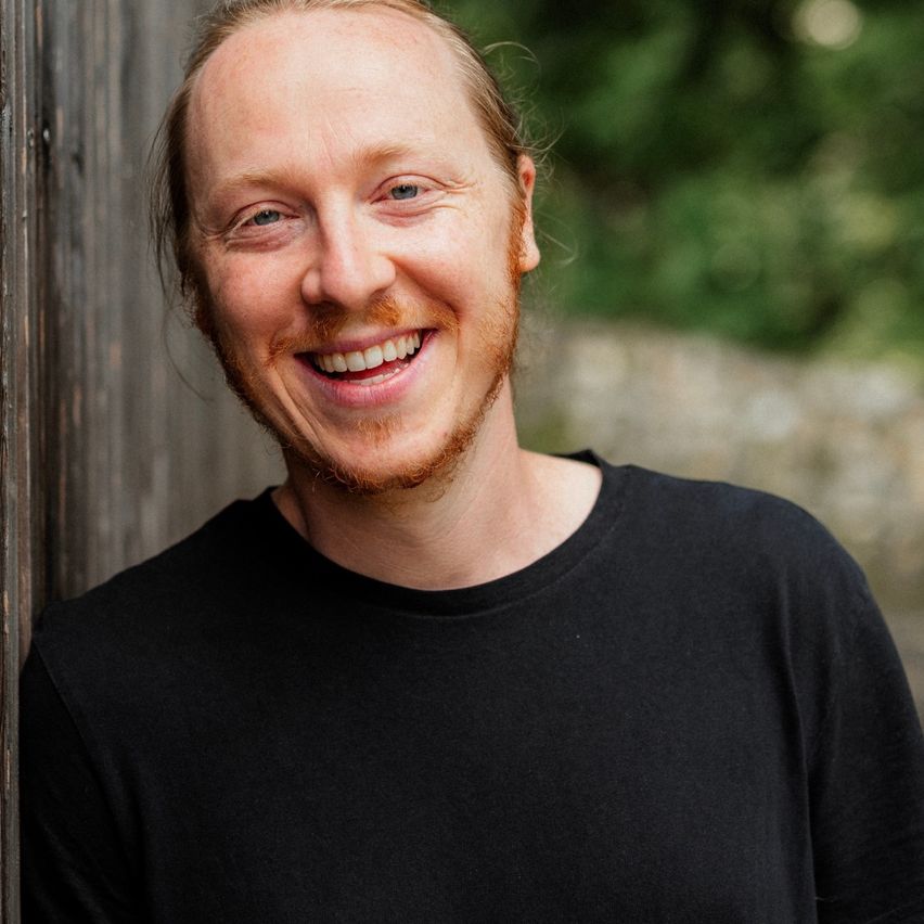 A smiling man with a beard is standing against a wooden wall. He is wearing a black t-shirt.