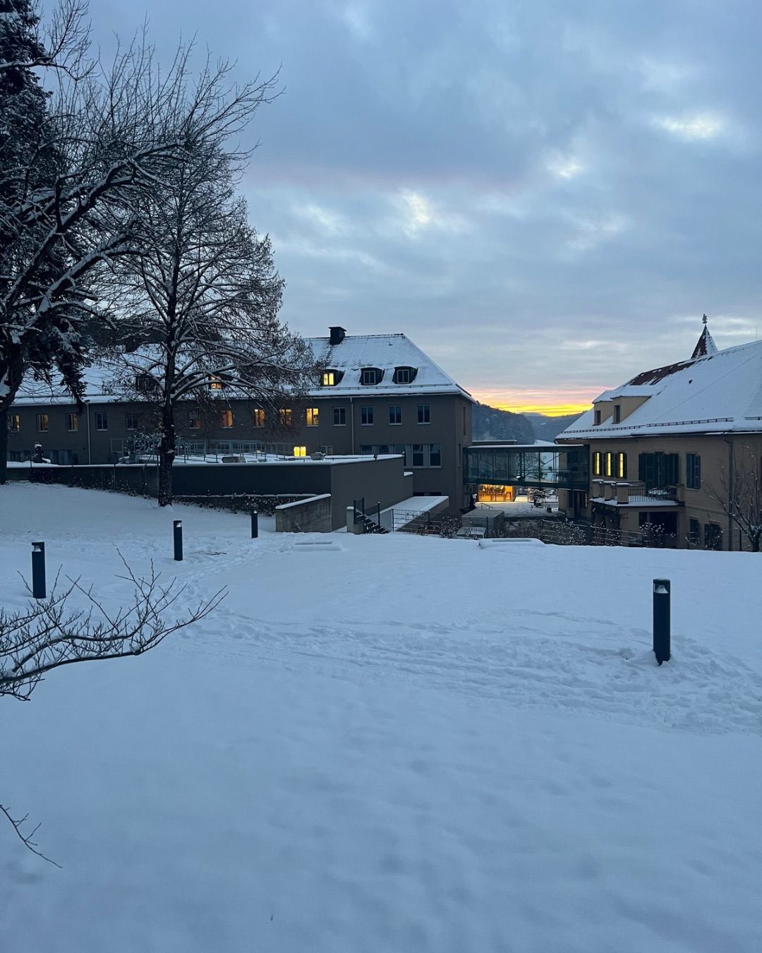 Eine Winterlandschaft zeigt ein Gebäude mit einer Glasbrücke, schneebedecktem Dach und warmen Lichtern. Der Himmel ist bewölkt und in der Ferne ist ein Blick auf Berge zu sehen.