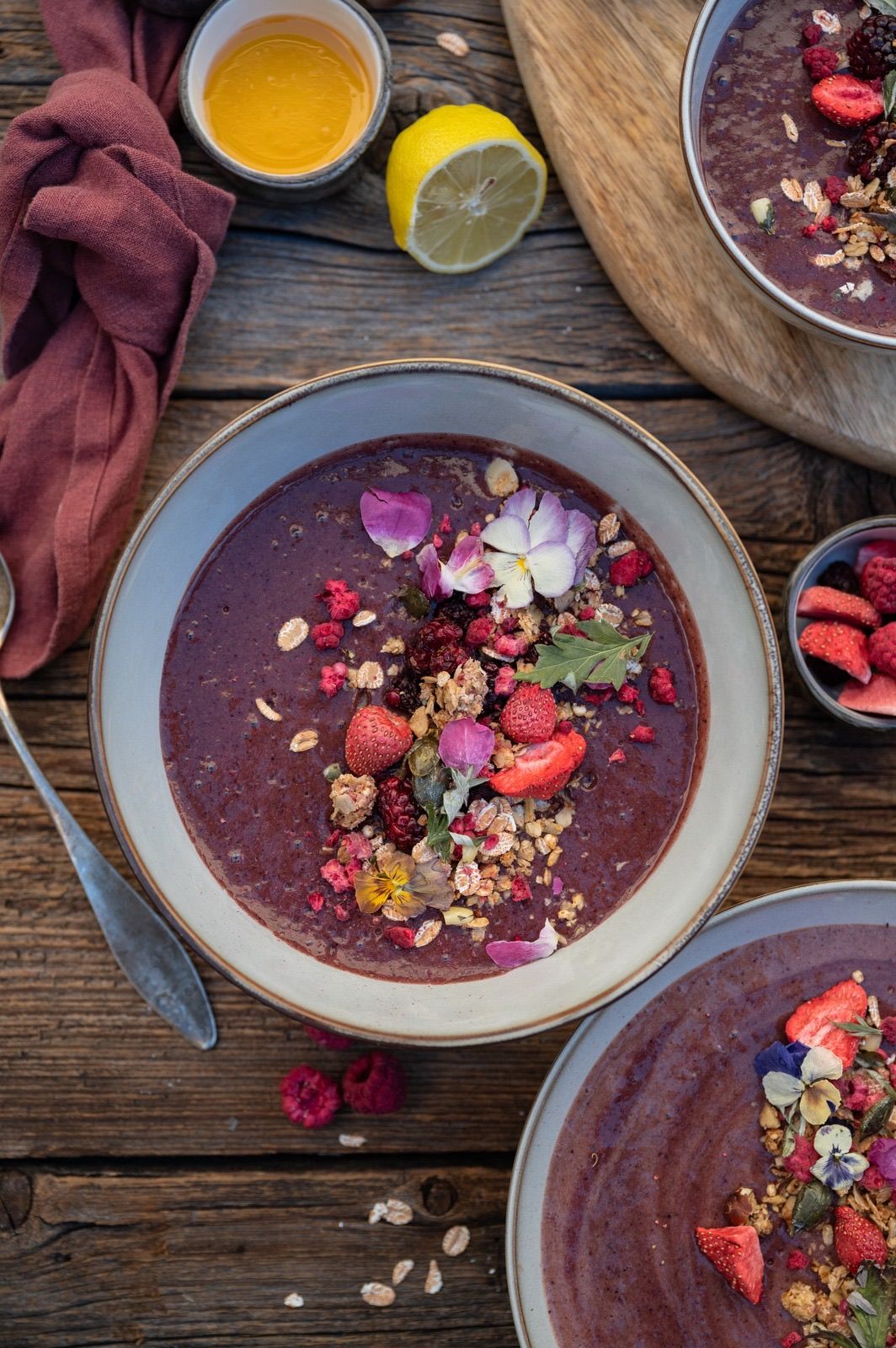 Eine Schüssel mit reichhaltigem lila Smoothie, garniert mit frischen Beeren, Blumen und Granola. Ein Löffel liegt in der Nähe, und eine Zitrone ist im Hintergrund sichtbar.
