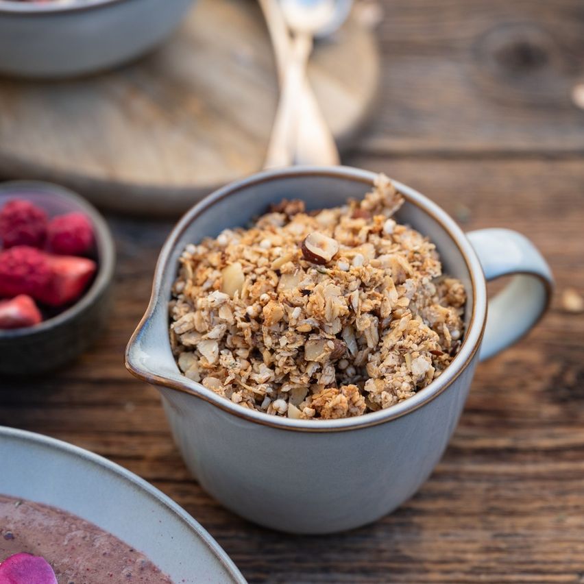 Eine Schüssel mit Granola steht auf einem Holztisch mit anderen Lebensmitteln. Eine kleine Schüssel hält rote Beeren und ein Holzteller hat zwei Löffel. Die Umgebung ist rustikal.