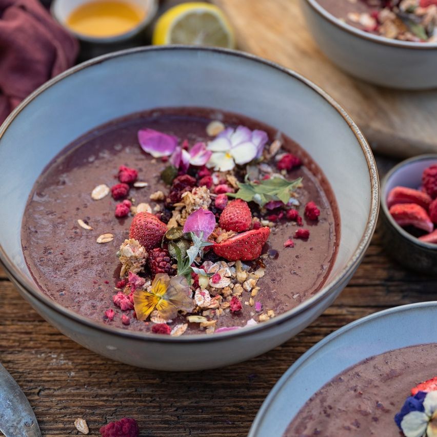 Eine Schüssel mit Schokoladen-Smoothie, garniert mit Erdbeeren, Himbeeren und Blumen auf einem Holztisch. Zusätzliche Schüsseln mit Honig und Zitrone befinden sich in der Nähe.