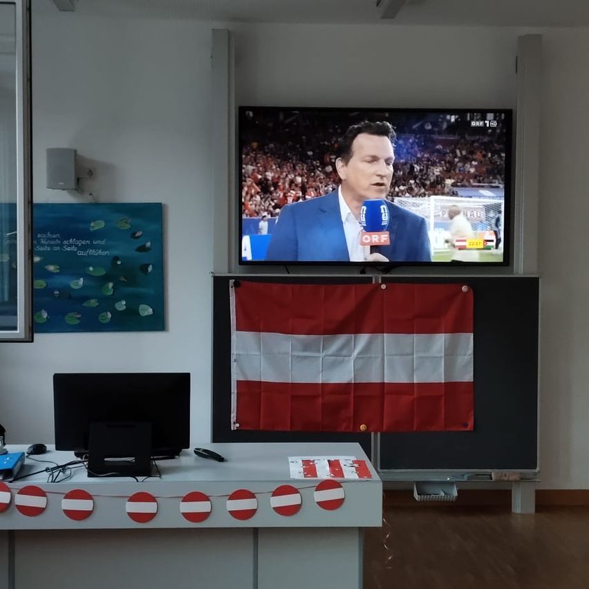 Ein Büro mit einem Fernsehbildschirm, der eine Sportübertragung zeigt, einer Flagge, einem Computerbildschirm und einer rot-weißen Banner auf dem Schreibtisch.