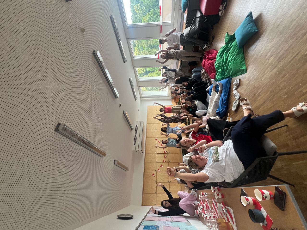 Eine Gruppe von Menschen in einem Raum, einige liegen auf dem Boden, andere sitzen in Stühlen, alle in Freizeitkleidung. Ein Tisch im Vordergrund hat Becher und Gläser.