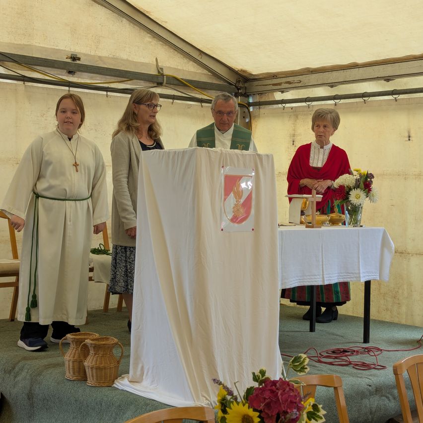 Eine Gruppe von Menschen, gekleidet in Weiß und Grün, steht hinter einem abgedeckten Altar in einem Zelt. Ein Mann in einer grünen Weste steht am Altar, mit Blumen und Körben in der Nähe.