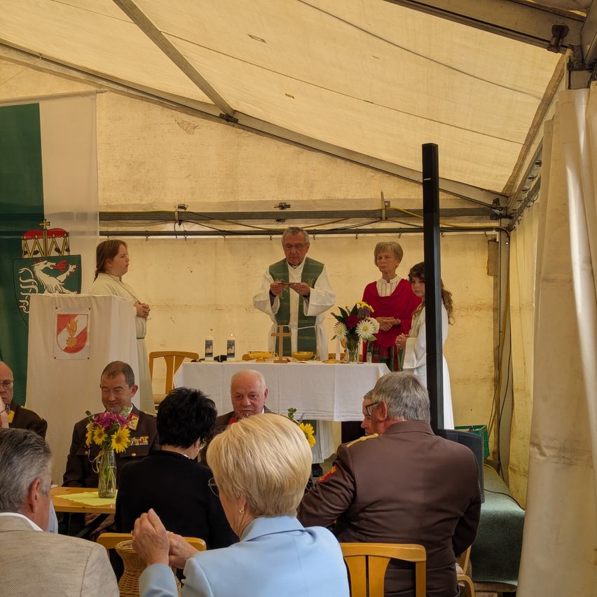 Eine Gruppe von Menschen in formeller Kleidung versammelt sich unter einem Zelt, wo ein Priester vor einem Tisch mit Blumen und Kerzen steht. Ein Banner mit einem Wappen ist ausgestellt.