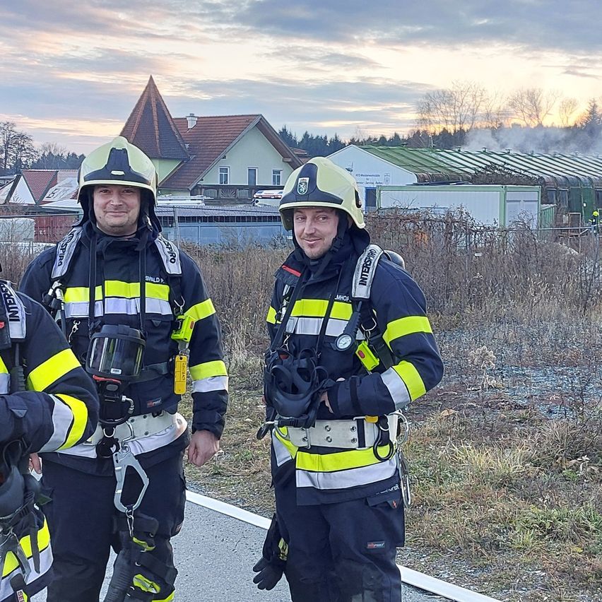 Drei Feuerwehrleute stehen am Straßenrand vor einem Gebäude, aus dem Rauch aufsteigt.