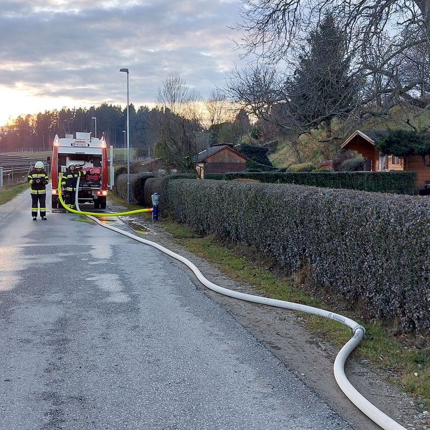 Zwei Feuerwehrleute stehen neben einem roten Feuerwehrwagen auf einer nassen Straße. Ein langer Schlauch erstreckt sich vom Wagen zu einem nahen Busch. Bäume und Häuser sind im Hintergrund zu sehen.