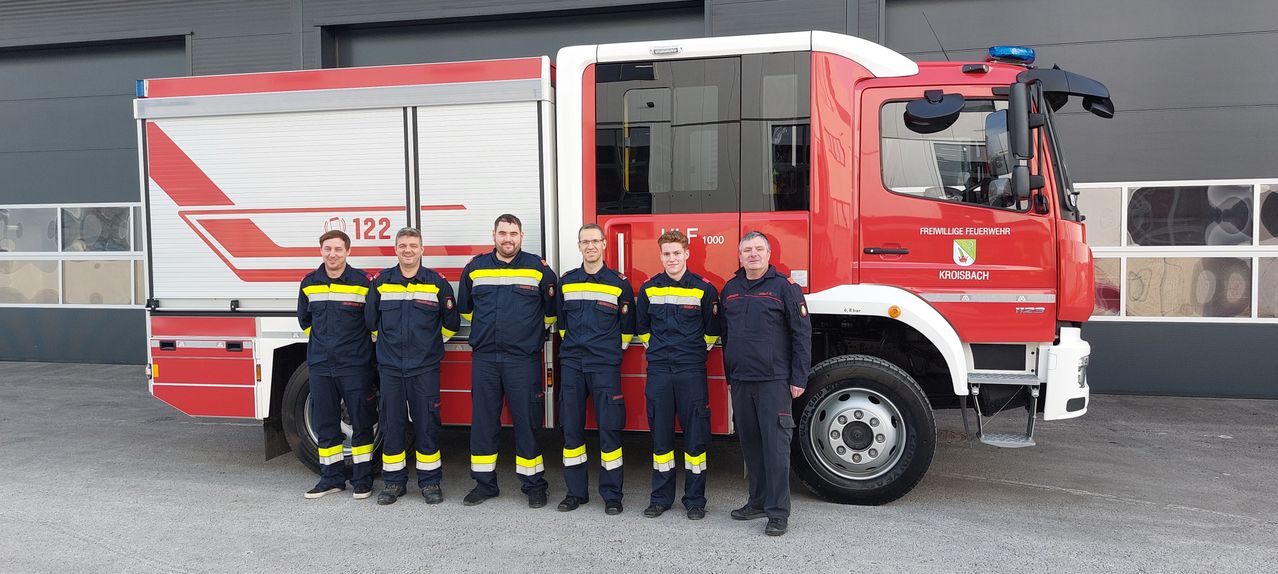 Fünf Feuerwehrleute stehen vor einem roten Feuerwehrwagen. Sie tragen Uniformen mit reflektierenden gelben und weißen Streifen. Die Tür des Fahrzeugs ist geöffnet, und die Nummer 122 ist auf der Seite angebracht.