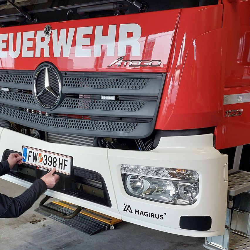 Eine Person hält das Nummernschild eines roten Mercedes-Benz-Lkws. Das Nummernschild lautet FW 398 HF. Der Lkw hat eine schwarze Kühlergrill mit dem Wort Magirus unter den Scheinwerfern.