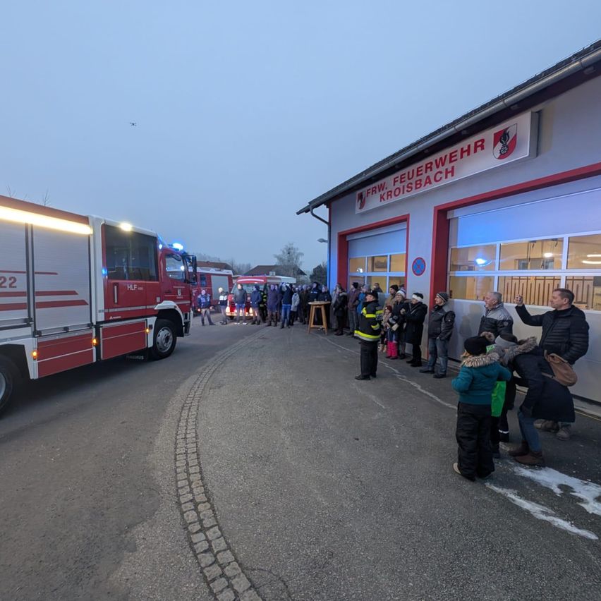 Ein roter Feuerwehrwagen steht vor der Freiwilligen Feuerwehr Kroisbach. Viele Menschen, einschließlich Kinder, sind vor dem Wagen versammelt.