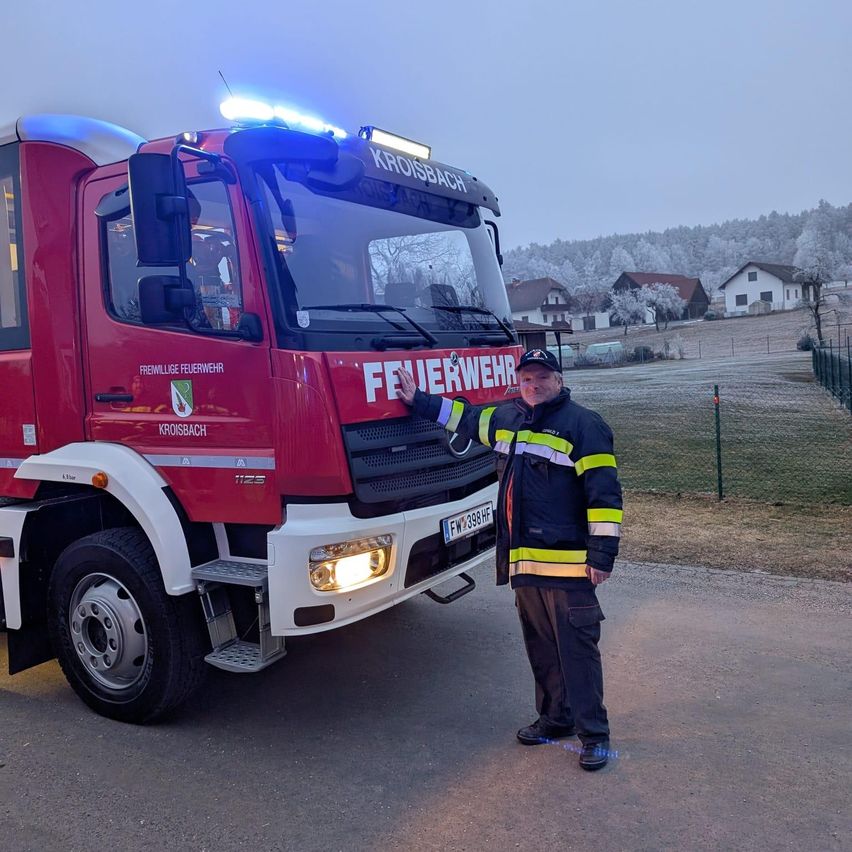Ein Feuerwehrmann steht neben einem roten Feuerwehrauto und zeigt auf es, mit einem Feld und Häusern im Hintergrund.