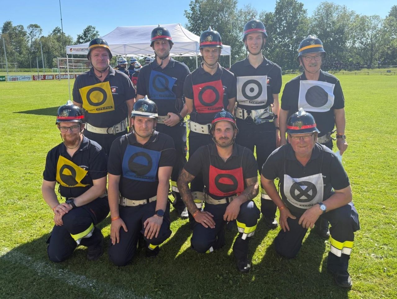 Eine Gruppe Feuerwehrleute posiert für ein Foto auf einem Rasenfeld, trägt Helme, Brillen und Uniformen mit kreisförmigen Symbolen. Hinter ihnen ist ein weißes Zelt zu sehen, und Bäume sind im Hintergrund.