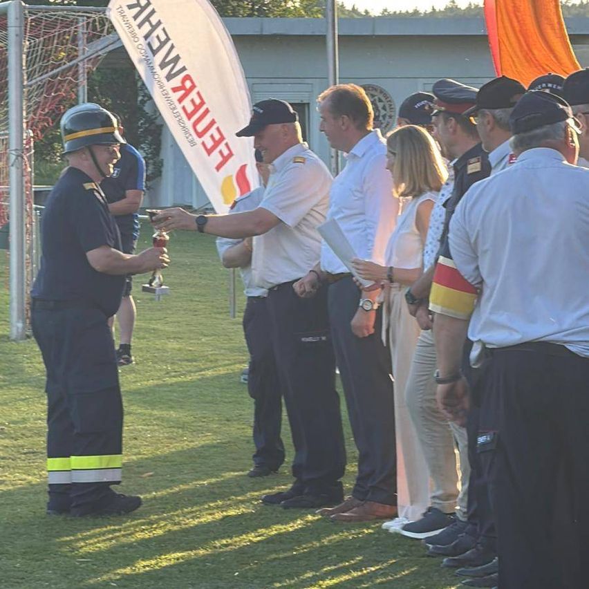 Eine Gruppe von Personen in Uniform steht auf einem Feld. Ein Mann mit Helm erhält eine Auszeichnung von einem anderen Mann in einer Kappe. Dahinter stehen weitere Personen in formeller Kleidung. Ein weißes Banner mit rotem Text ist sichtbar.