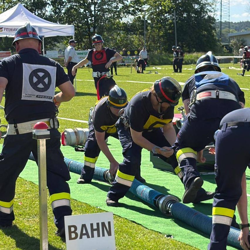 Feuerwehrleute nehmen an einer Trainingsübung auf einem Feld teil. Eine Person beugt sich über einen Schlauch. Ein Schild mit dem Wort Bahn ist in der Nähe.