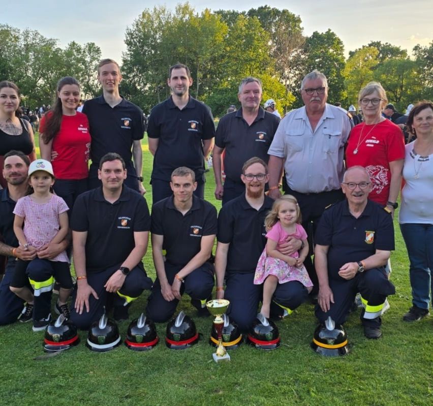 Eine Gruppe Feuerwehrleute und ihre Familien posieren auf einem Rasenfeld für ein Foto, mit Pokalen und Helmen vor ihnen.