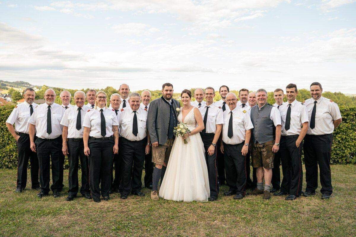 A wedding party with the bride and groom standing in the center, surrounded by a group of men in matching uniforms.