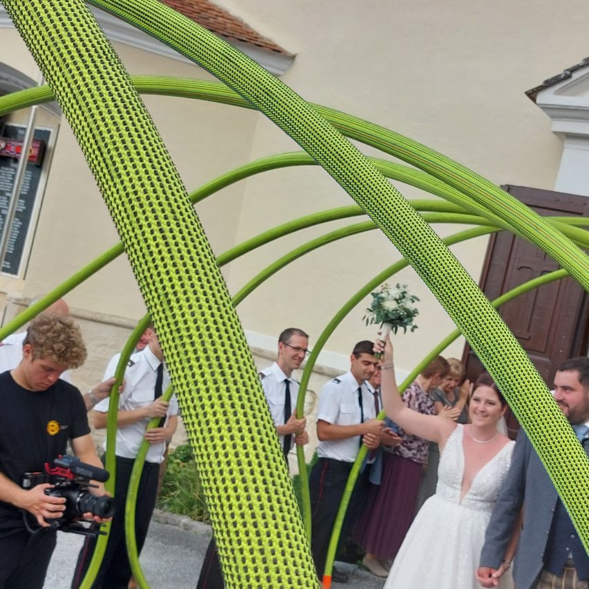 A bride in a white dress holds a bouquet of flowers under a green arch, with people around her, including a man holding a camera.
