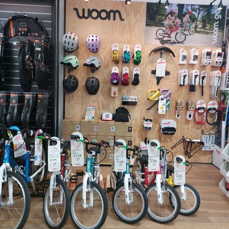 Fahrradgeschäft mit verschiedenen Fahrrädern, Helmen, Handschuhen und Zubehör. Ein großes Schild mit dem Wort 'wOom' ist an der Wand angebracht. Ein Plakat eines Kindes, das Fahrrad fährt, ist an der Wand montiert.