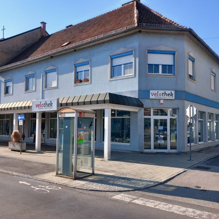 Ein blaues Gebäude mit einem braunen Dach und blauem Rand hat mehrere Fenster und ein Schild mit der Aufschrift Velothek. Ein Telefonhäuschen steht auf dem Bürgersteig.