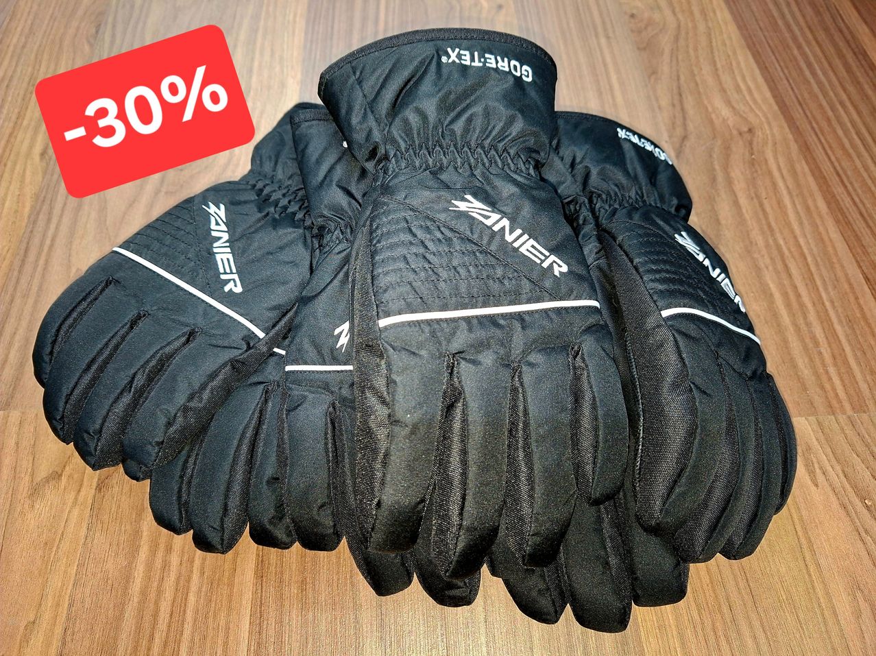 Ein Paar schwarze Winterhandschuhe mit dem Wort Vanier und Gore-Tex werden auf einer Holzoberfläche ausgestellt. Ein rotes Schild mit dem Text 30% ist auf der linken Seite platziert.