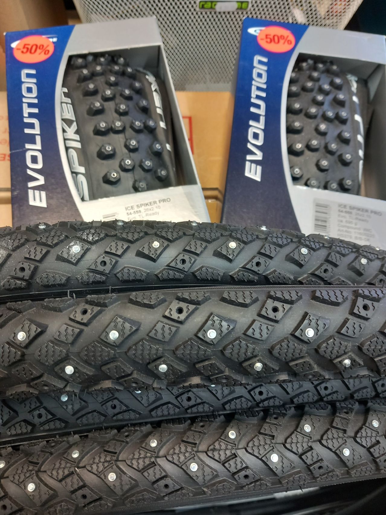 Zwei Paar Evolution Spiken Ice Spiker Pro Reifen, die aufeinandergestapelt sind, mit Kartons im Hintergrund.