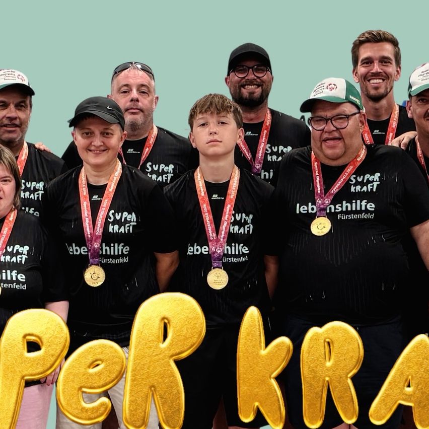 Eine Gruppe von Menschen in gleichen schwarzen T-Shirts und Medaillen, mit 'Super Kraft' auf der Vorderseite. Sie stehen zusammen vor einem türkisen Hintergrund mit goldenen Buchstaben, die 'Super Kraft' lesen.