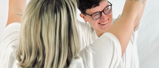 A woman with short hair and glasses smiles while another person with long hair looks on.