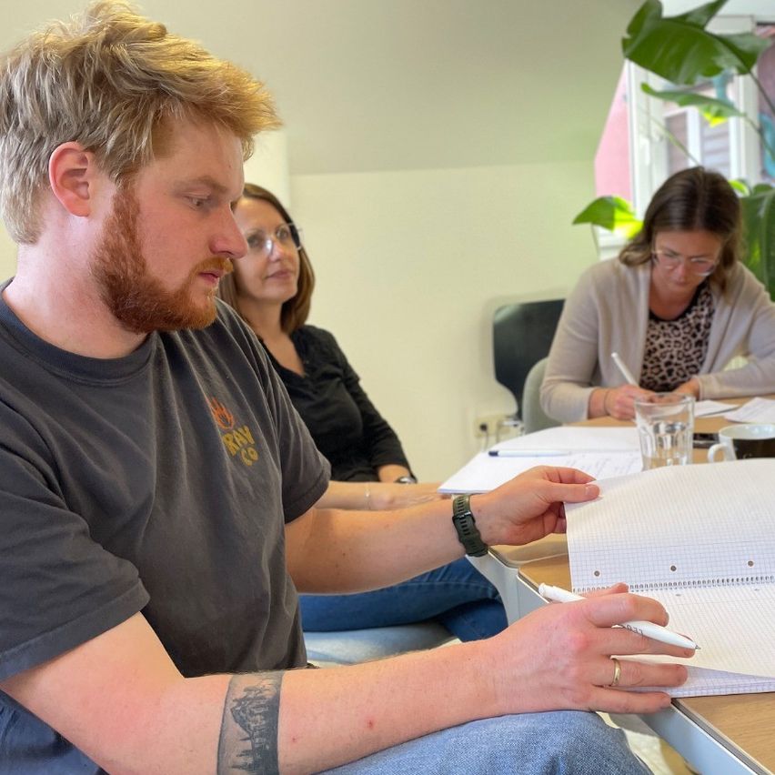 Drei Menschen sitzen an einem Tisch, zwei Frauen und ein Mann. Der Mann hält einen Stift und Papier und scheint in eine Diskussion mit den Frauen verwickelt zu sein.