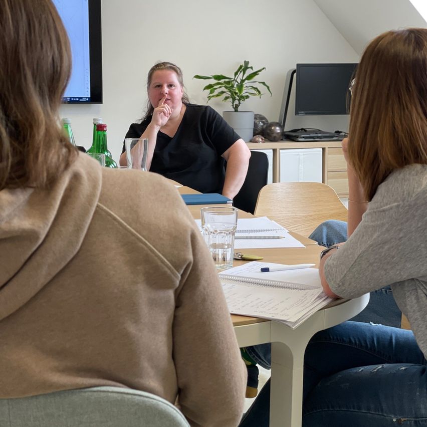 Drei Frauen sitzen in einem Besprechungsraum an einem Tisch. Eine von ihnen spricht mit der Hand am Kinn.