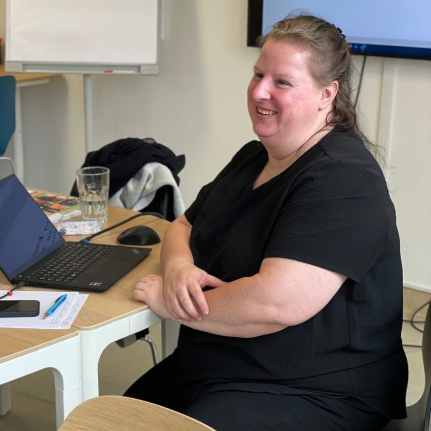 Eine Frau in einem schwarzen Shirt sitzt an einem Schreibtisch in einem Büro. Sie lächelt und schaut nach vorne. Der Schreibtisch hat einen Laptop, einen Stift, ein Handy und ein Glas Wasser. Ein Whiteboard ist an der Wand hinter ihr befestigt.
