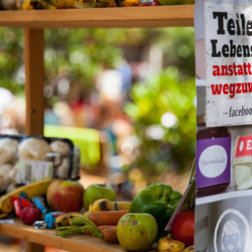 Ein Holzregal mit verschiedenen Obst- und Gemüsesorten, Bananen, Äpfeln, grünen Paprikaschoten und Karotten. Ein Schild mit dem Text 'Teile Lebens anstatt wegzu' ist ebenfalls zu sehen.
