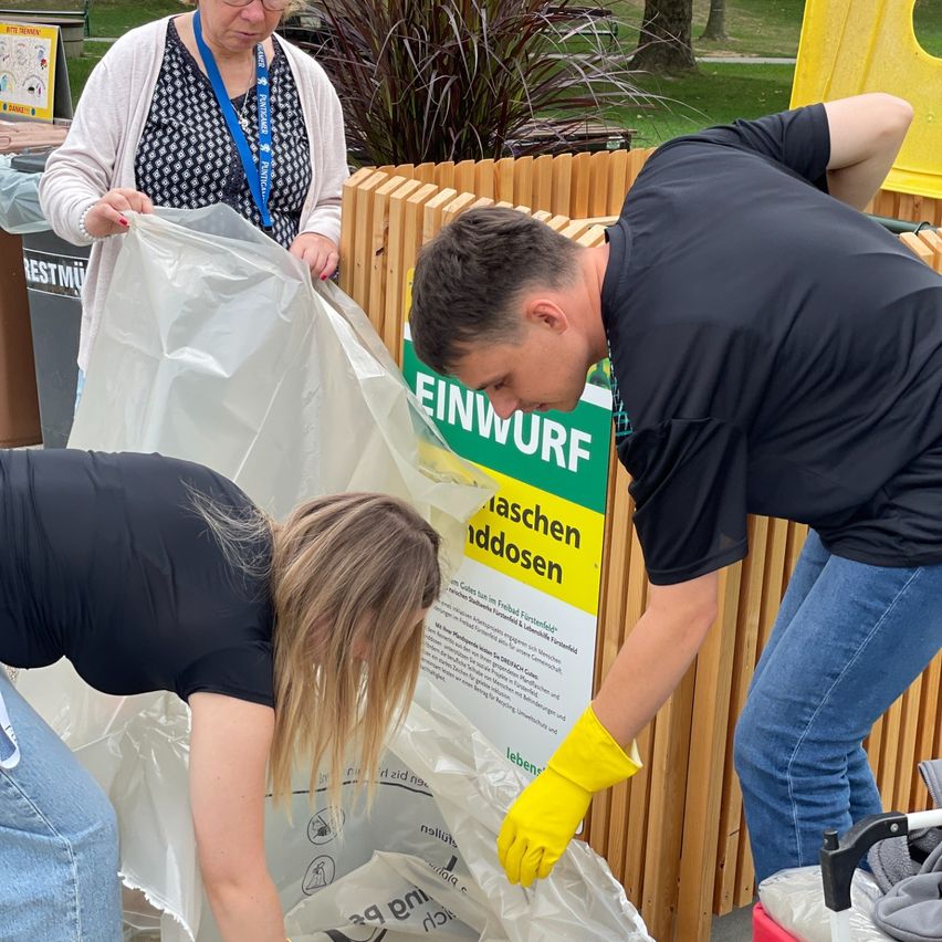Drei Personen sortieren Müll in einen durchsichtigen Plastiksack. Der Mann trägt gelbe Handschuhe. Hinter ihnen befindet sich ein Schild mit dem Wort 'Einwurf'.