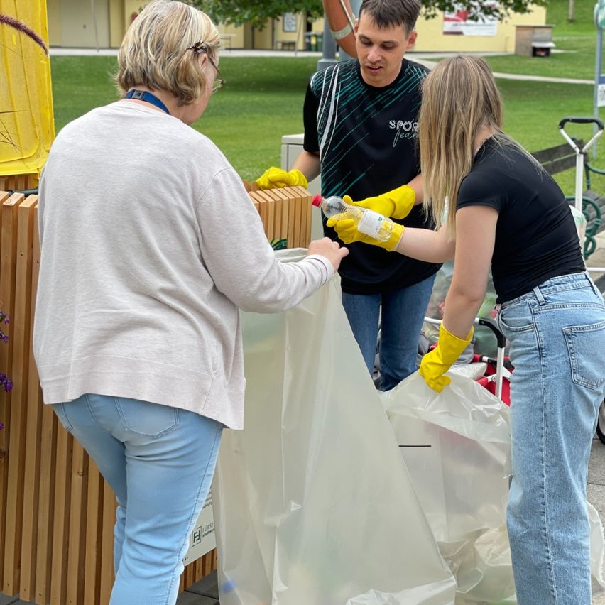 Drei Personen reinigen einen Außenbereich und tragen Handschuhe. Eine Frau hält eine Plastiktüte, eine andere Frau eine Flasche und ein Mann steht hinter ihnen.