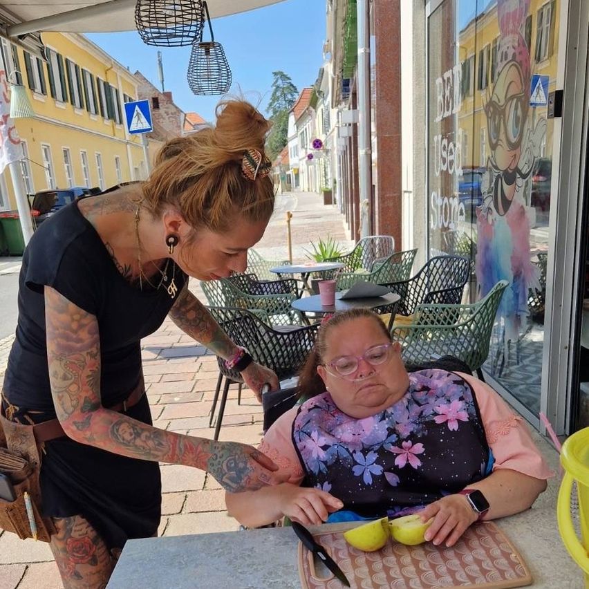 Eine Frau mit Tattoos hilft einer Frau mit Brille, Äpfel auf einem Tisch vor einem Café zu schneiden.