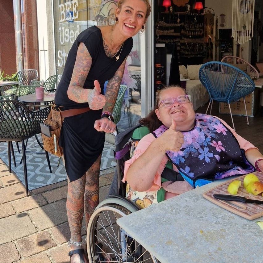 Zwei Frauen sind draußen in der Nähe eines Restaurants, eine im Rollstuhl, beide zeigen Daumen hoch. Sie haben Obst auf einem Tisch vor sich.