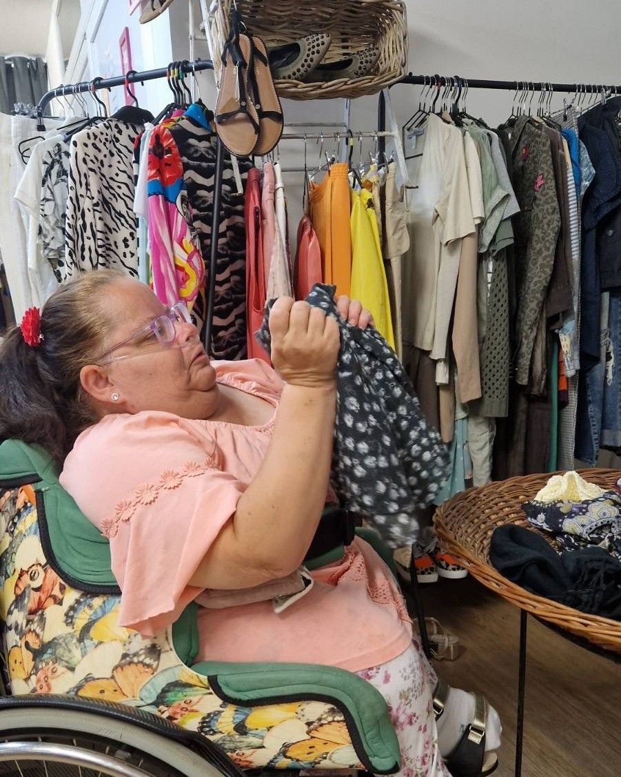 Eine Frau in einem rosa Blusen sitzt auf einem Stuhl und untersucht ein Kleidungsstück. Hinter ihr steht ein Kleiderständer mit verschiedenen Kleidungsstücken. Ein Korb mit Kleidung steht auf dem Boden.