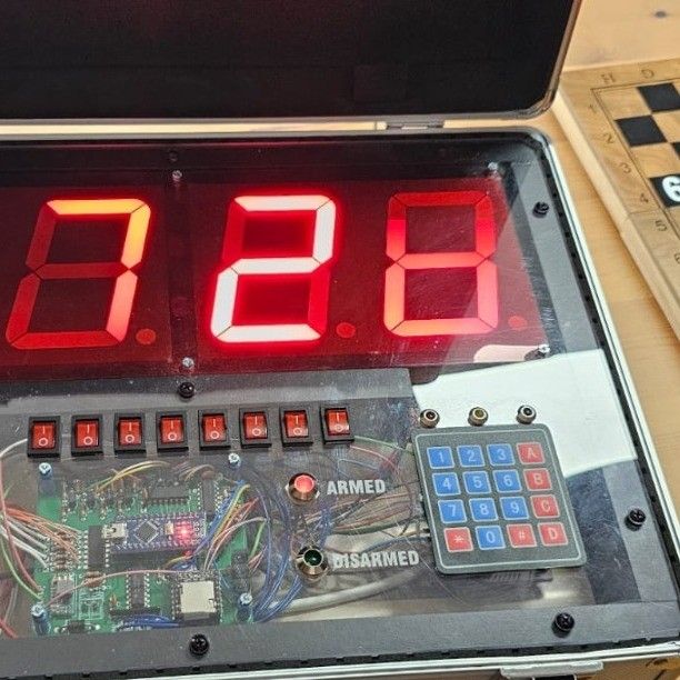 Ein elektronisches Gerät mit einem digitalen Display, das die Zahl 828 zeigt. Unter dem Display befinden sich rote Tasten und eine Tastatur mit Buchstaben und Zahlen. Links davon ein komplexes Leiterplatten-Design mit verschiedenen Kabeln.