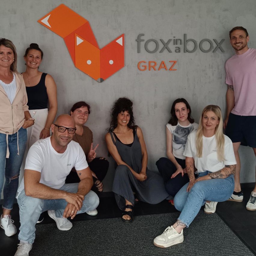 Eine Gruppe von Menschen posiert für ein Foto vor einem Fox in Box Graz Logo an einer Wand. Sie lächeln und sitzen oder knien auf dem Boden.