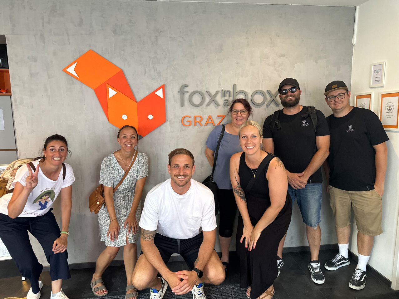 Eine Gruppe von sieben Personen steht lächelnd vor einer fox@box GRAZ Wand. Ein Mann in Weiß kniet, und eine Frau in Schwarz steht neben ihm.