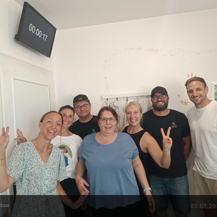 Eine Gruppe lächelnder Erwachsener steht in einem Raum. Die Wand hinter ihnen ist weiß gestrichen. Eine digitale Uhr über der Tür zeigt 00:017 an.